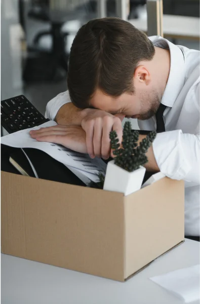 An overwhelmed employee rests his head on his arms over a cardboard box filled with papers and a small potted plant on a desk.