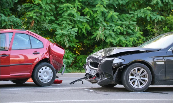 A red car with rear-end damage and a black car with front-end damage are parked closely on a road surrounded by lush greenery.