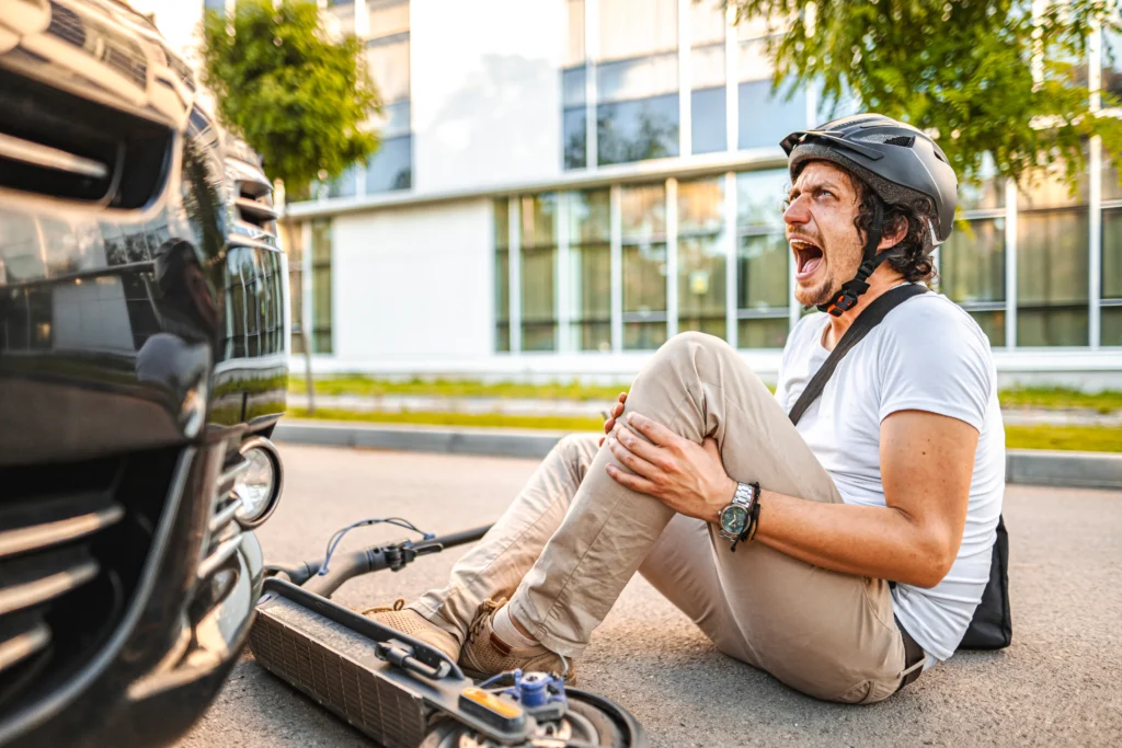 An injured person seated on the road with a scooter beside them, looking concerned as a black car approaches.