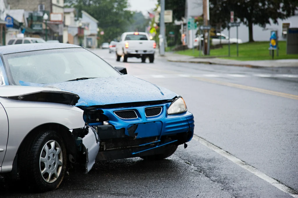 Blue car with significant front damage sits in the rain beside a silver car, amidst a quiet street scene.