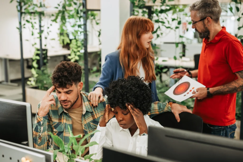 A group of colleagues in a modern office appears stressed, with two individuals sitting at a desk, surrounded by lush greenery.
