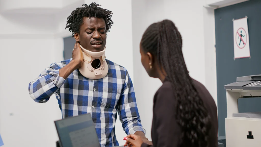 person wearing a training device for CPR stands facing a woman engaged in conversation, with office equipment in the background.