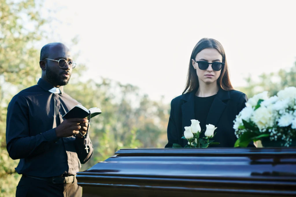 Solemn outdoor funeral scene featuring a coffin adorned with flowers, as a clergy member reads from a book.