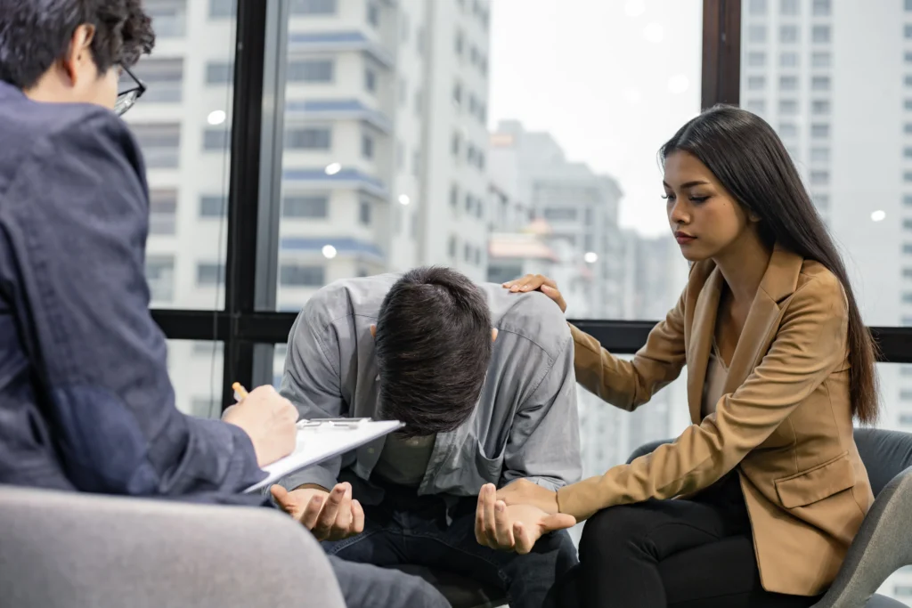 Man leans forward in distress, while a woman comforts him, as an observer takes notes in a modern, urban counseling setting.