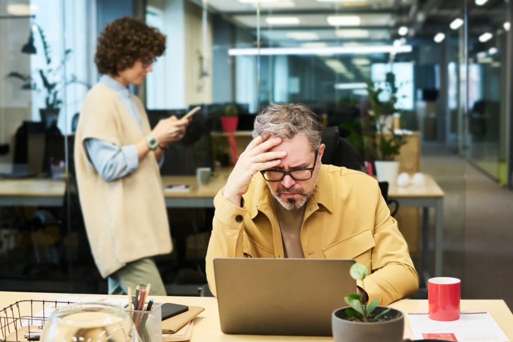 A man in a yellow jacket looks stressed while working on a laptop, surrounded by plants and office supplies in a modern workspace.