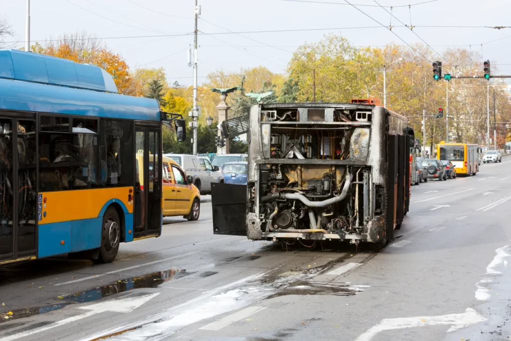 Burnt-out bus reveals its charred interior on a busy street, while a blue bus and cars are visible in the background.