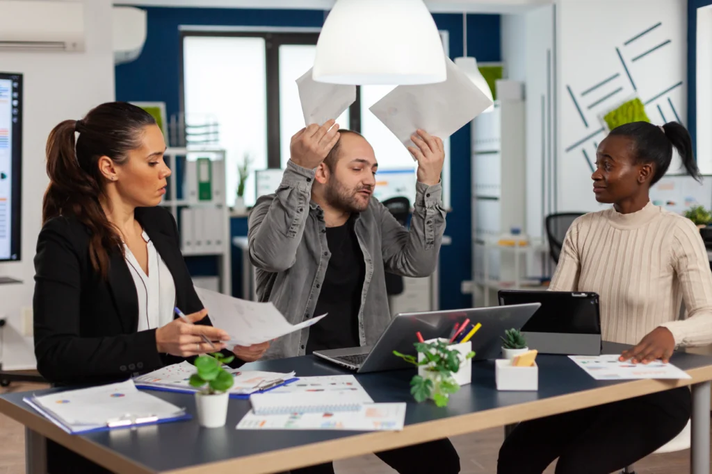 A tense office meeting with one person gesturing animatedly while holding documents, as colleagues observe around a table.