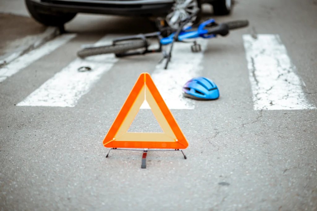 An orange warning triangle on a crosswalk near a fallen bicycle and a blue helmet, indicating a recent accident.