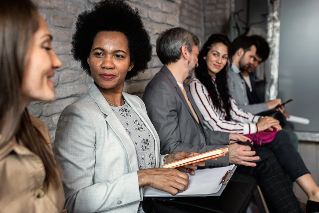 Five people sit in a row on chairs against a brick wall, holding notebooks and papers.