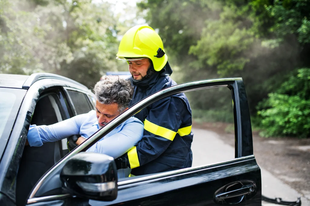 A firefighter in a bright yellow helmet assists an injured person from a car, surrounded by greenery and smoke.