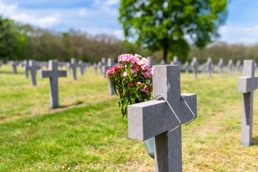 A cemetery filled with gray crosses, with a flower pot of pink blooms attached to one, under a blue sky and green trees.