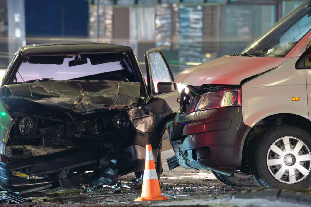 Two damaged cars in a night-time accident scene, with a traffic cone marking the site of collision and debris scattered around.