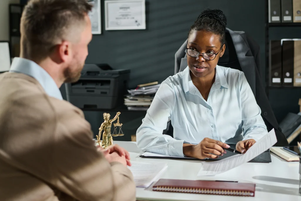 A professional meeting at an office desk, featuring a woman reviewing documents, with a statue of Justice and organized files in the background.