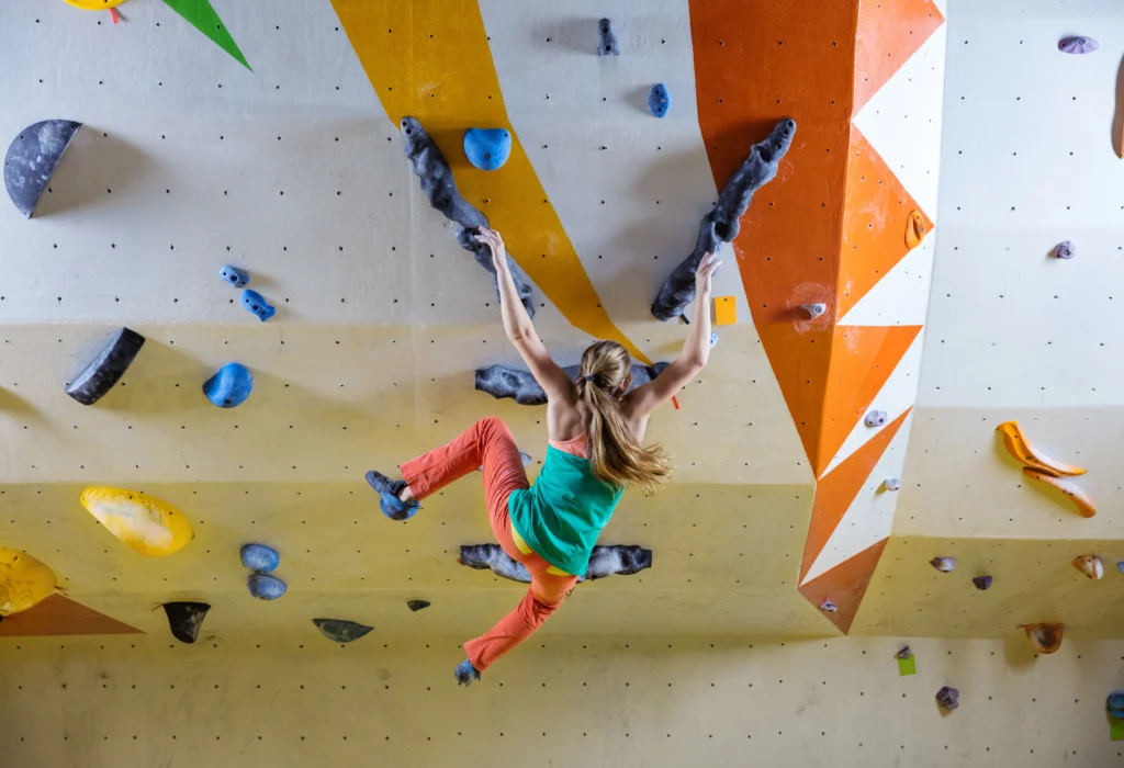 Climber in a green top and orange pants maneuvers on an indoor climbing wall adorned with colorful holds.