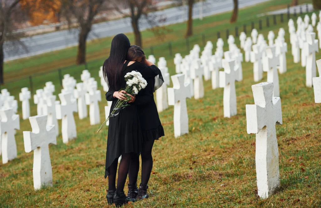 Two individuals in black attire embrace while holding white flowers, surrounded by rows of white gravestones in a serene cemetery.