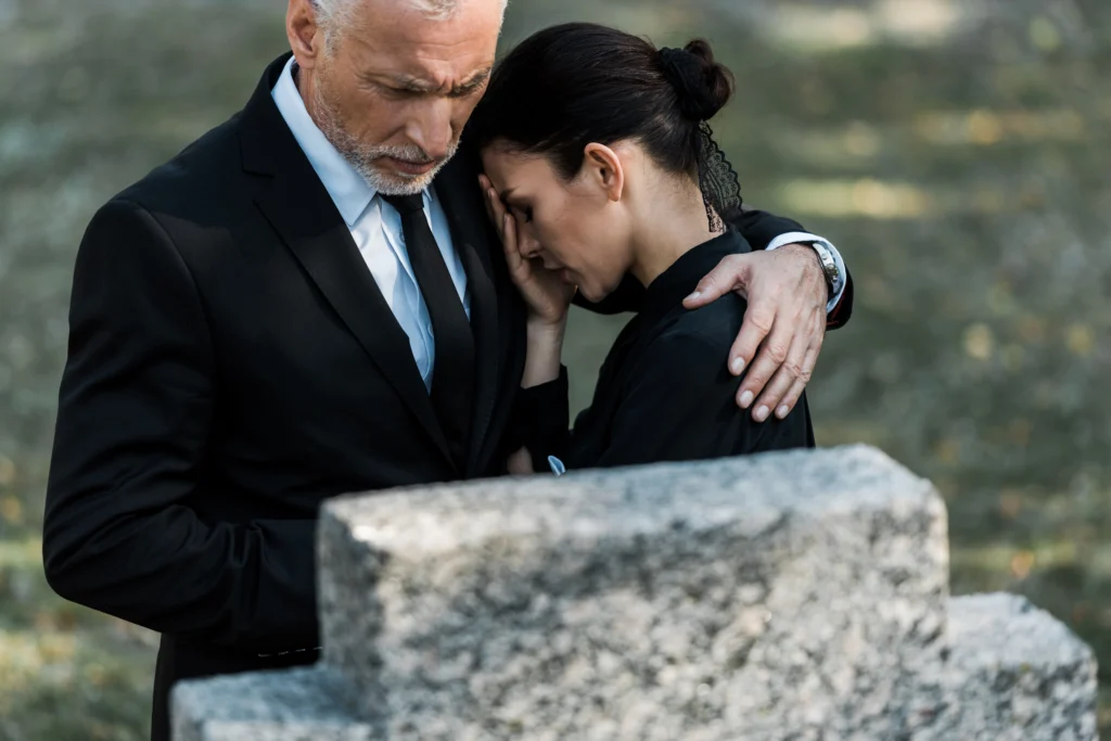 A man in a black suit comforts a woman in black attire at a grave, suggesting a moment of grief and support between them.