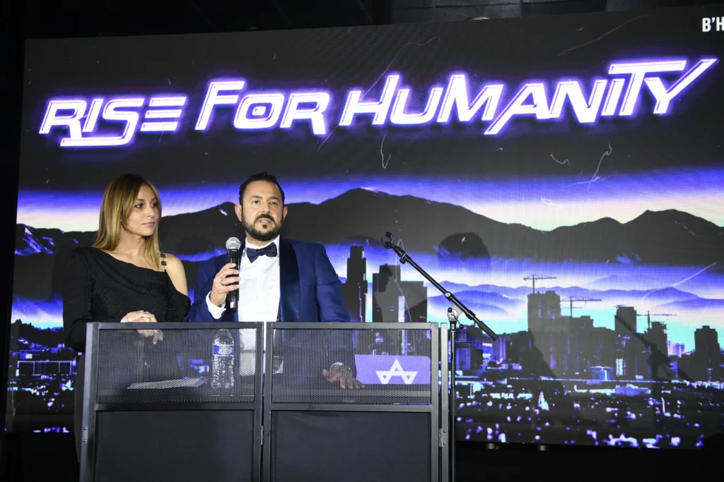 A Alex tofer speaks at a podium, while a woman in a black dress stands beside him, with a cityscape backdrop and "Rise For Humanity" displayed.